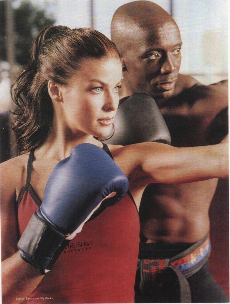 Carmen Electra and Tae Bo creator Billy Blanks on the February 1999 cover of Muscle & Fitness magazine, both wearing Carushka Bodywear. Carmen wears the signature red Mary Jane Tank Top with the Carushka logo.