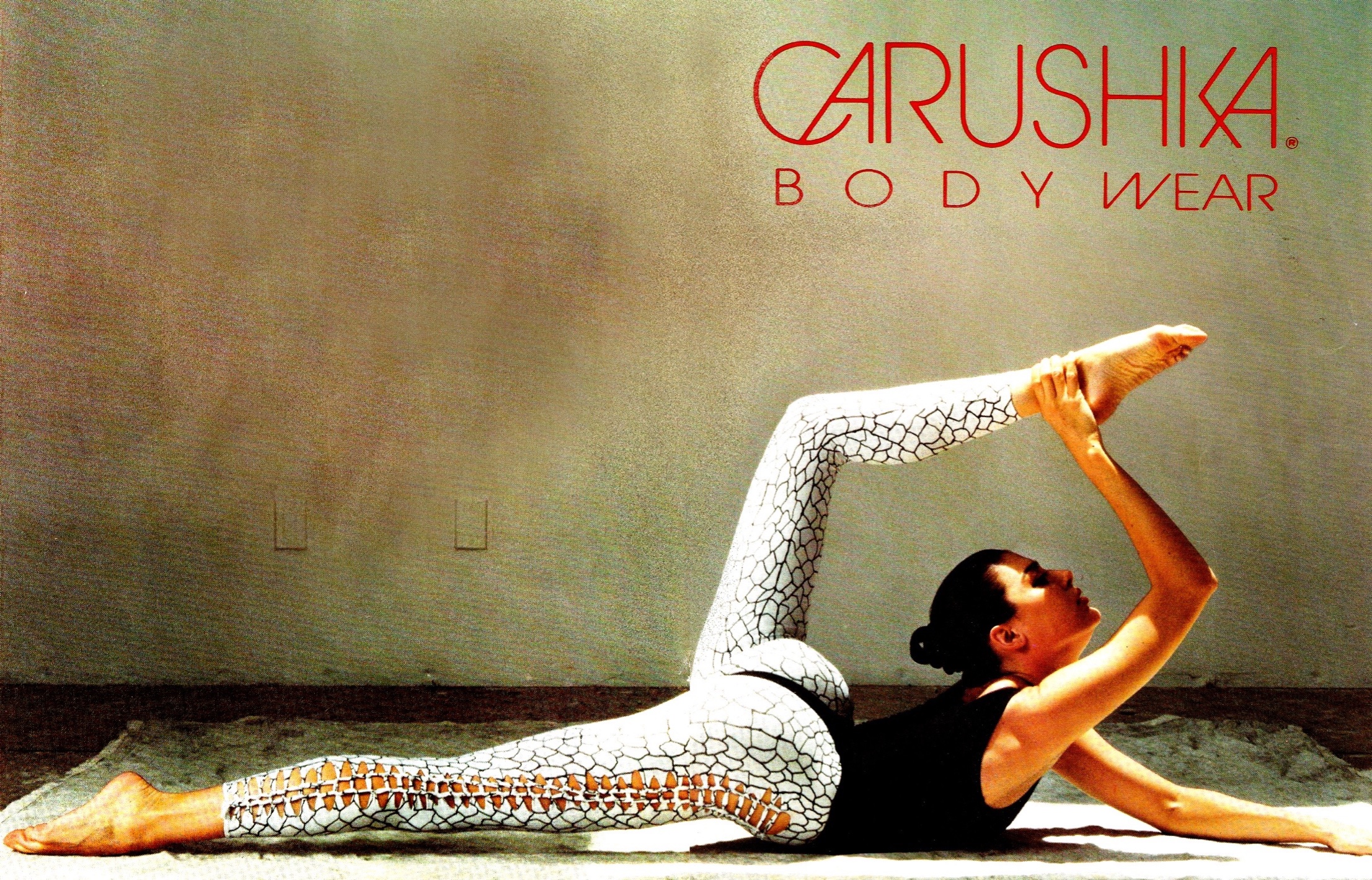 A dramatic fashion pose with a dancer holding her foot over her head featuring Carushka's 1990 signature lizard-print webb ankle tight leggings in white and a classic high-rise black thong-back tank leotard, highlighting her architectural approach to unique bodywear design.  The original designs that started yoga wear and athleisure
