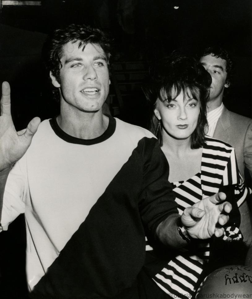 Fashion designer Carushka Jarecka and actor John Travolta posing together to promote 'Travolta by Carushka,' their collaborative 1983 menswear and bodywear line launched in conjunction with the film Staying Alive. Carushka is seen in a signature striped ensemble, representing the brand's Hollywood era.
